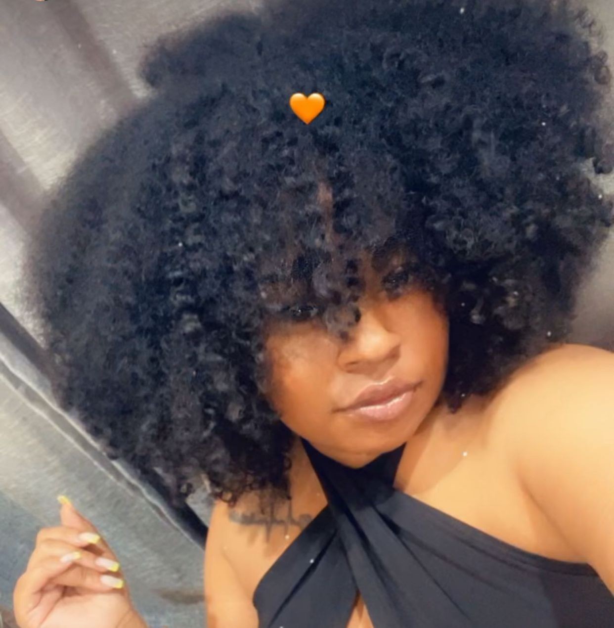 Person with curly hair and an orange heart emoji above their head, wearing a black outfit.