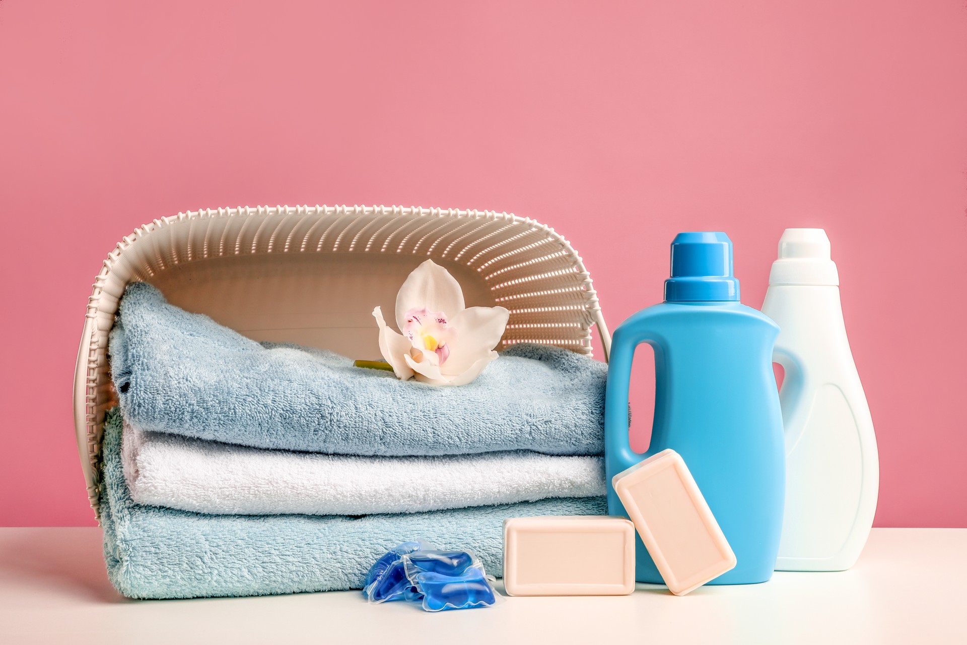 Stack of bath towels with white orchid flower in laundry basket, detergents, washing capsules and soap on pink background