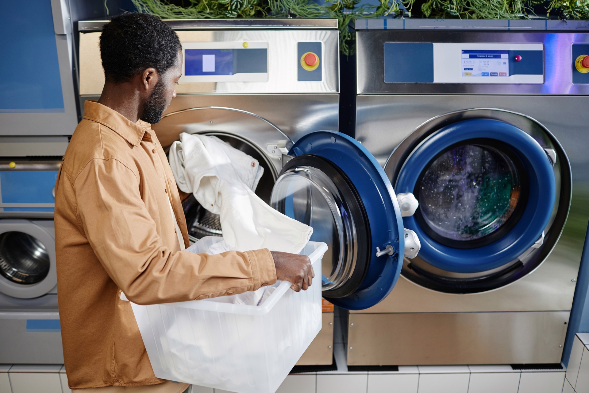 Side view of young black man in beige shirt putting white clothes into machine